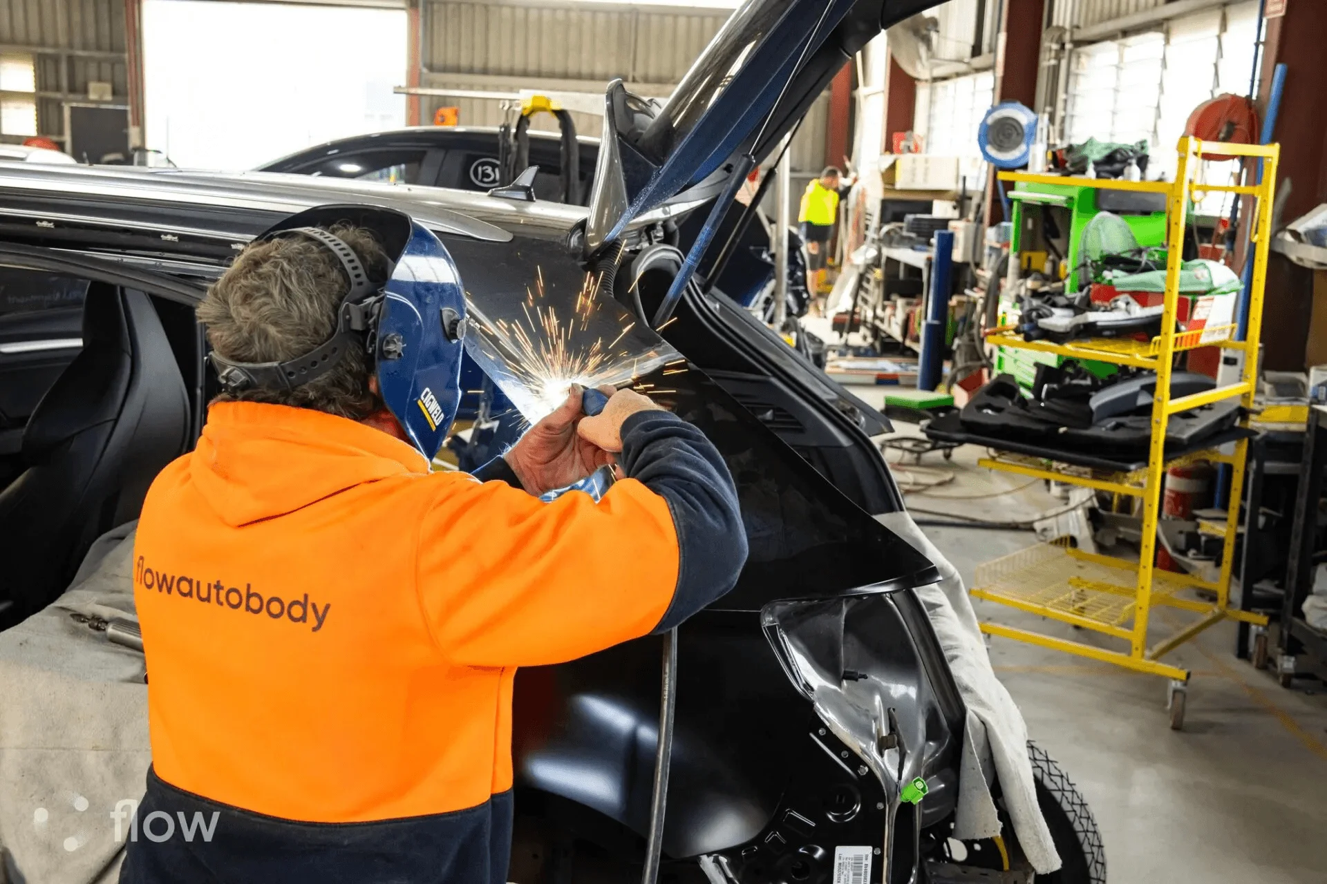 A Flow Autobody technician welding a vehicle, with the Flow Autobody name across the back of his orange work shirt